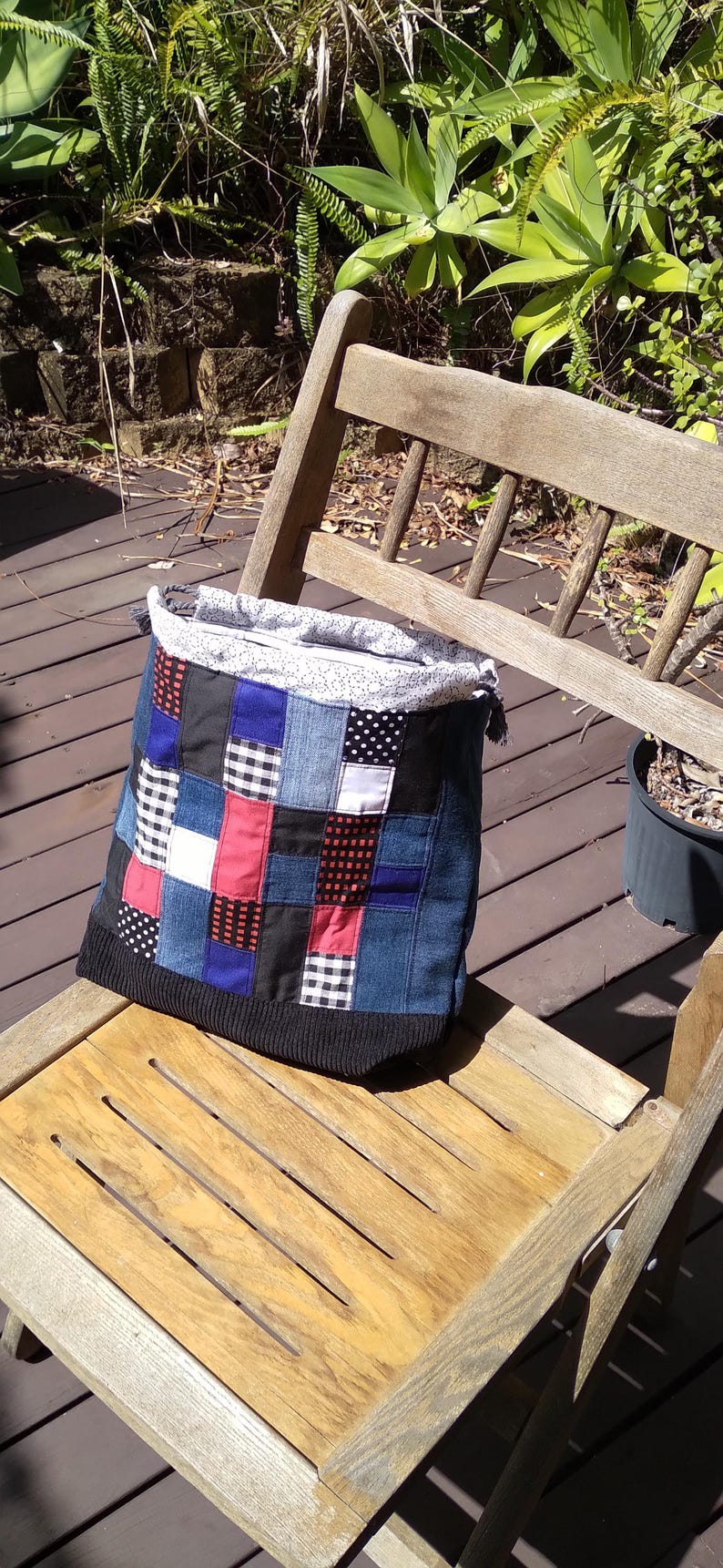 May include: A patchwork tote bag with a denim body and a black knit bottom sits on a wooden chair. The patchwork design includes squares of blue denim, red, black, and white patterned fabrics. The bag's top is a light-colored fabric with a small white pattern.