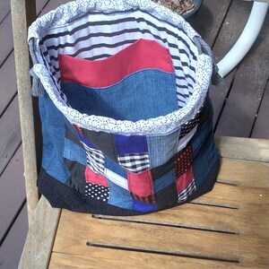 May include: A handmade patchwork tote bag with a denim exterior. The bag features a variety of fabric patches in blue, red, black, and white. The interior lining is striped, and the top edge is trimmed with white lace.