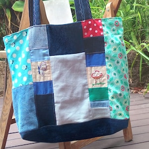 Puede incluir: Un bolso tote patchwork con paneles de mezclilla y tela estampada. La bolsa presenta detalles bordados de una mariposa y un hongo. La bolsa tiene asas de mezclilla y se exhibe en una silla de madera.