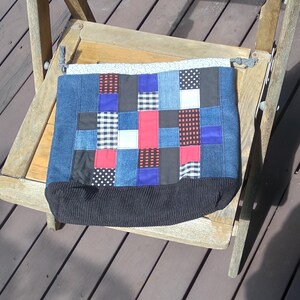May include: A patchwork tote bag with a denim base and a variety of fabric squares in red, blue, black, and white patterns. The bag has a black ribbed bottom and is resting on a wooden chair.