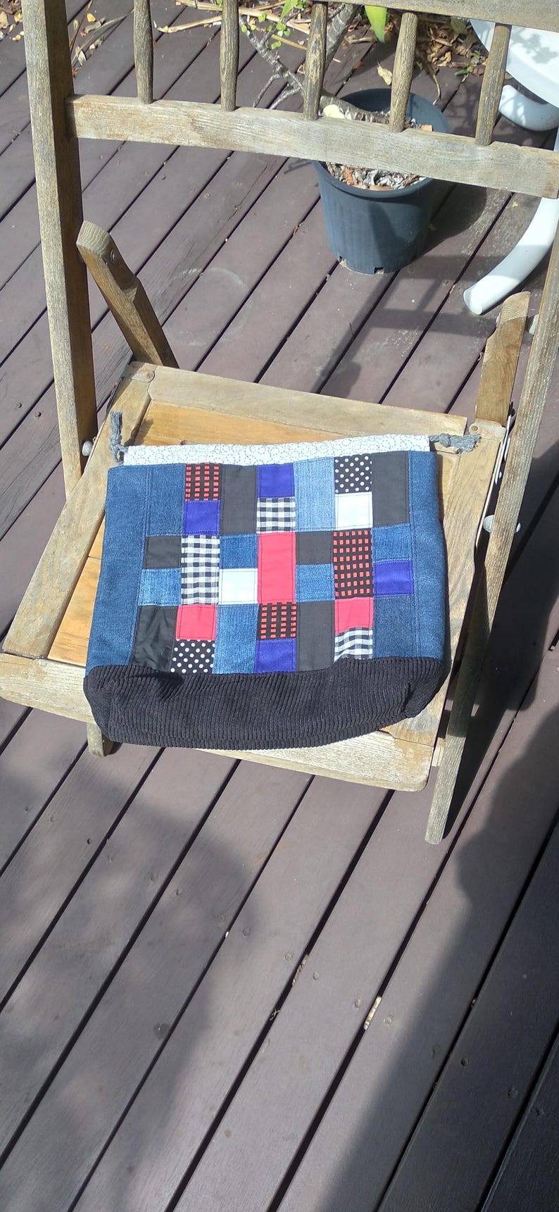 May include: A patchwork tote bag with a denim base and a variety of fabric squares in red, blue, black, and white patterns. The bag has a black ribbed bottom and is resting on a wooden chair.