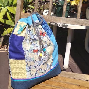 Puede incluir: Una bolsa con cordón de patchwork con base de mezclilla y una variedad de telas, incluyendo floral, azul y ganchillo amarillo. Adornada con aplicaciones de mariposas y pájaros, y un cordón gris.