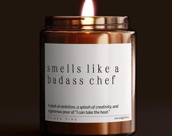 May include: A lit candle in a brown glass jar with a white label. The label reads "smells like a badass chef." Below the text is a quote and the brand name "GILDED PINE." The candle weighs 8 oz.