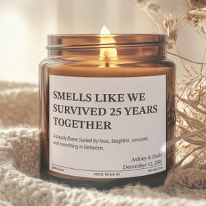 A brown glass candle labeled "Smells like we survived 25 years together" sits on a white knitted blanket, with a dried flower arrangement in the background.