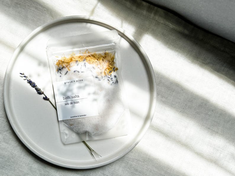 A white plate with a package of bath salt on it, placed on a white surface with sunlight shining on it.