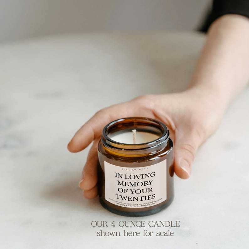 A hand holding a brown candle labeled "In Loving Memory of Your Twenties" on a white surface.