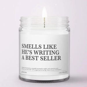 May include: A clear glass jar candle with a white label featuring the black text: SMELLS LIKE HE'S WRITING A BEST SELLER. Below this, smaller text reads: A dash of inspiration, a sprinkle of sleepless nights, and a generous pour of just one more edit—all wrapped up in one big cheers to your masterpiece. The candle has a lit wick.