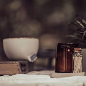 a table with various objects, including a brown candle, a white bowl, a brown jar, a brown bottle, a brown vase, and a brown book. The objects are arranged in a casual and relaxed setting, with the focus on the table and its contents.