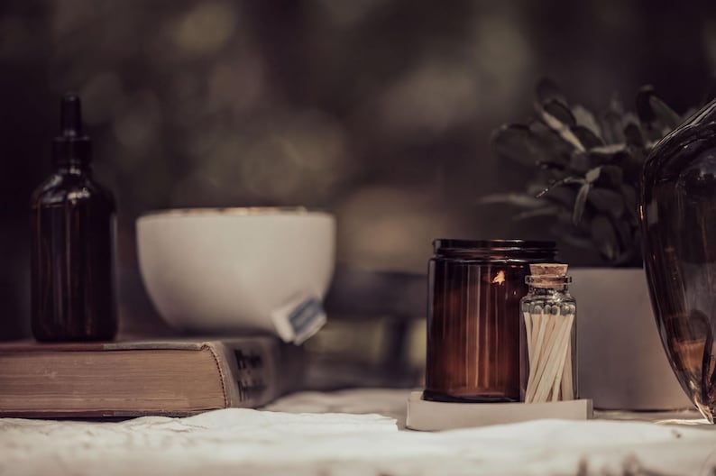 a table with various objects, including a brown candle, a white bowl, a brown jar, a brown bottle, a brown vase, and a brown book. The objects are arranged in a casual and relaxed setting, with the focus on the table and its contents.