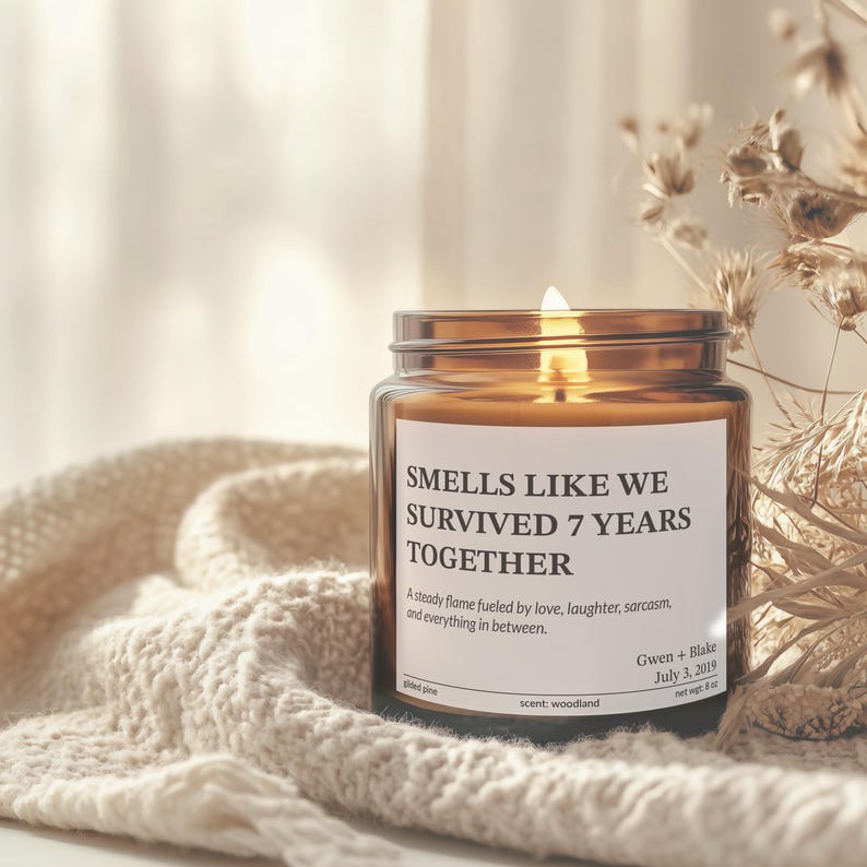 A brown candle labeled "Smells like we survived 7 years together" sits on a white knitted blanket, with a blurred background.