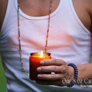 A person is holding a lit candle in their hand, wearing a white tank top and a colorful beaded necklace.