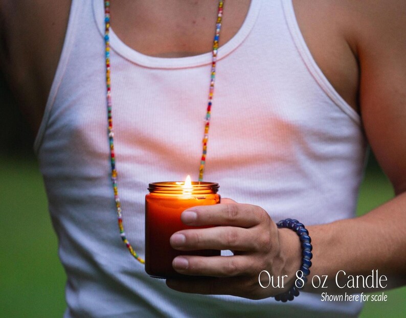 A person is holding a lit candle in their hand, wearing a white tank top and a colorful beaded necklace.
