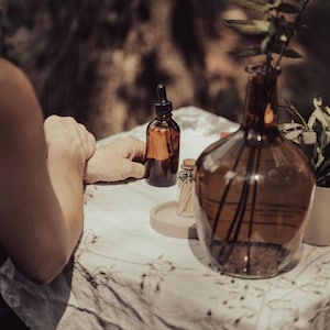 A person's hands are resting on a table with various objects, including a glass bottle, a glass vase with a plant, and a small glass container.