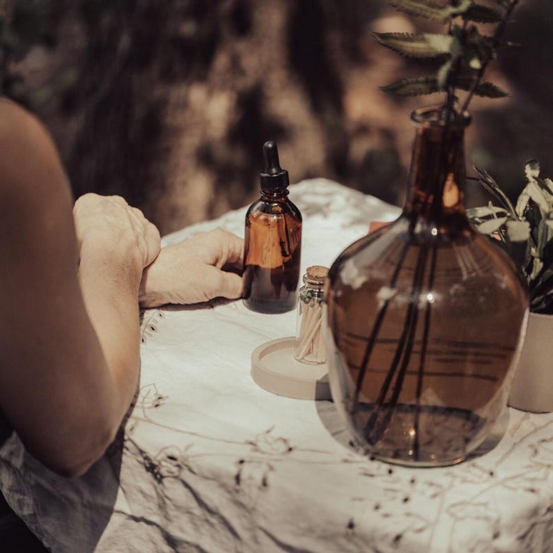A person's hands are resting on a table with various objects, including a glass bottle, a glass vase with a plant, and a small glass container.
