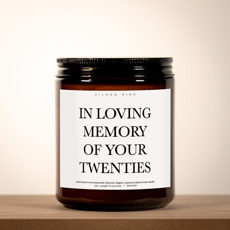 A brown glass jar with a black lid, featuring a white label with black text that reads "IN LOVING MEMORY OF YOUR TWENTIES".