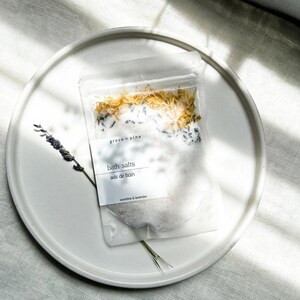 A white plate with a package of bath salt on it, placed on a white surface with sunlight shining on it.