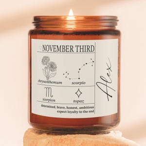 May include: A lit candle in an amber glass jar with a white label featuring the text NOVEMBER THIRD, a chrysanthemum illustration, a Scorpio constellation, a scorpion symbol, a topaz symbol, and the name Alex in script.