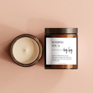 May include: A brown glass jar candle with a black lid and a white label that reads "SENDING YOU A - big hug". The candle is vanilla scented and has a cream-colored wax. The text on the label includes a personal message.