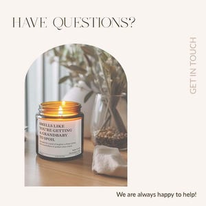 a candle labeled "Smells like you're getting older & grandbaby, get to spoil" on a wooden surface, with a vase of greenery and a towel nearby.
