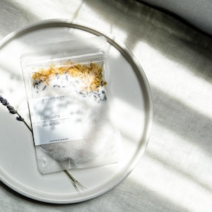 A white plate with a package of bath salt on it, placed on a white surface with sunlight shining on it.