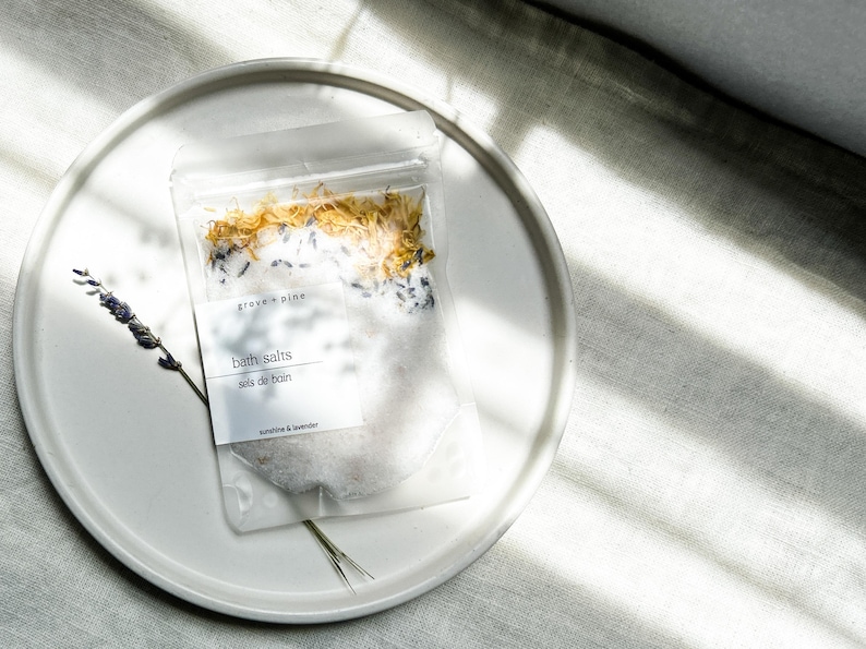A white plate with a package of bath salt on it, placed on a white surface with sunlight shining on it.