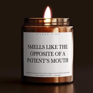 May include: A lit, amber-colored glass jar candle with a metal lid. The label reads "SMELLS LIKE THE OPPOSITE OF A PATIENT'S MOUTH." The candle is hand-poured with a natural vegan coconut blend wax and weighs 8 ounces.