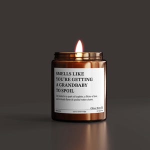 A brown candle with a lit wick sits on a dark surface, with a label that reads "SMELLS LIKE YOU'RE GETTING A GRANDBABY TO SPOIL".