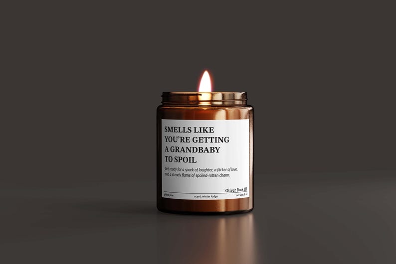 A brown candle with a lit wick sits on a dark surface, with a label that reads "SMELLS LIKE YOU'RE GETTING A GRANDBABY TO SPOIL".