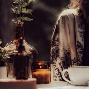 A woman is sitting at a table with a white mug, a candle, and a glass vase with a plant.