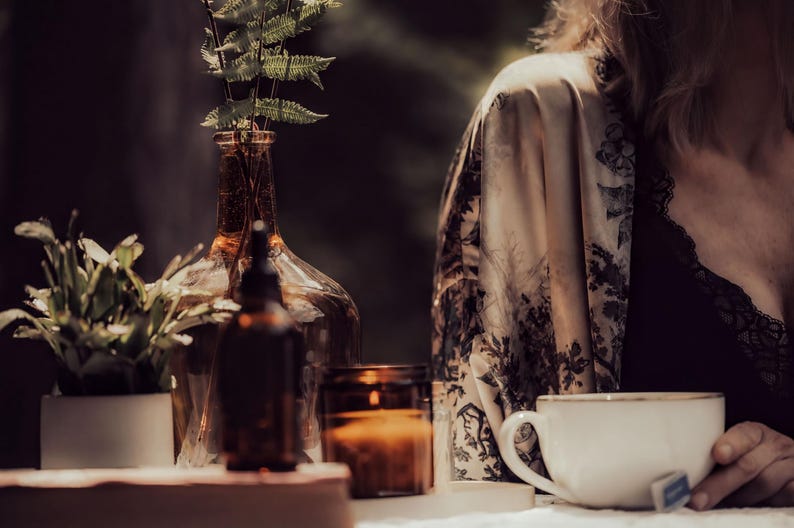A woman is sitting at a table with a white mug, a candle, and a glass vase with a plant.