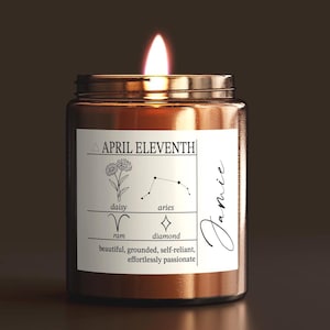 May include: A lit candle in a brown glass jar with a label that reads "APRIL ELEVENTH". The label includes a daisy illustration, the Aries constellation, and the words "ram" and "diamond". The candle is labeled "Jamie" and has the words "beautiful, grounded, self-reliant, effortlessly passionate".