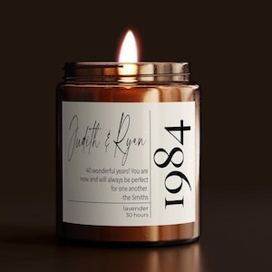May include: A brown glass jar candle with a lit wick and a white label. The label features the text Judith & Ryan, 1984, 40 wonderful years! You are now and will always be perfect for one another. -the Smiths, lavender, and 30 hours.