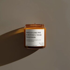 A brown glass jar candle with a white label that reads "SMELLS LIKE WE SURVIVED 1 YEAR TOGETHER" and includes a date and scent.
