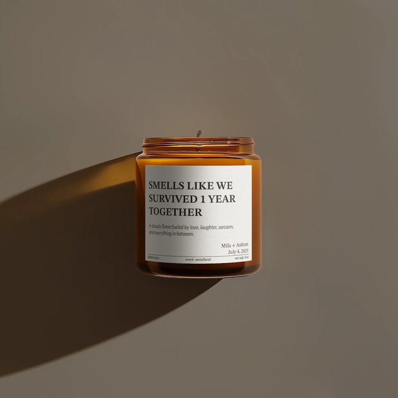 A brown glass jar candle with a white label that reads "SMELLS LIKE WE SURVIVED 1 YEAR TOGETHER" and includes a date and scent.