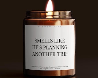 May include: A lit candle in a brown glass jar with a white label. The label reads "SMELLS LIKE HE'S PLANNING ANOTHER TRIP." The candle is a small batch, hand-poured, natural vegan coconut blend wax candle, net weight 8 ounces.