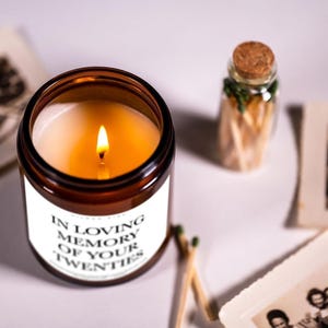 a candle labeled "In Loving Memory of Your Twenties" surrounded by a small glass jar with matches and a photograph.