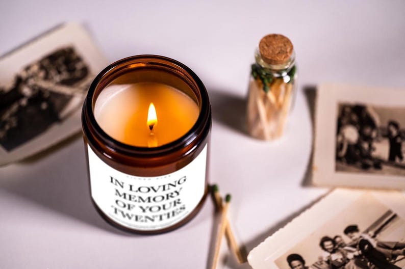 a candle labeled "In Loving Memory of Your Twenties" surrounded by a small glass jar with matches and a photograph.