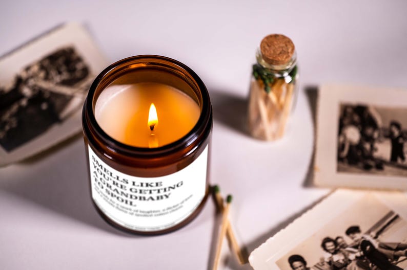 a brown candle with a lit flame, a small glass jar with a cork lid, and some photographs arranged on a white surface.