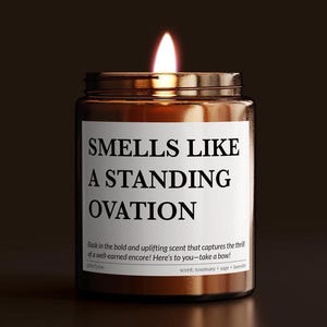 May include: A lit candle in a brown glass jar with a white label. The label reads "SMELLS LIKE A STANDING OVATION." The scent is rosemary, sage, and lavender. The candle is a home decor item.