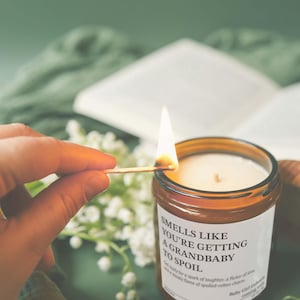 A hand is lighting a candle with a match, and the candle is labeled with the text "Smells like you're getting to spoil a grandbaby".