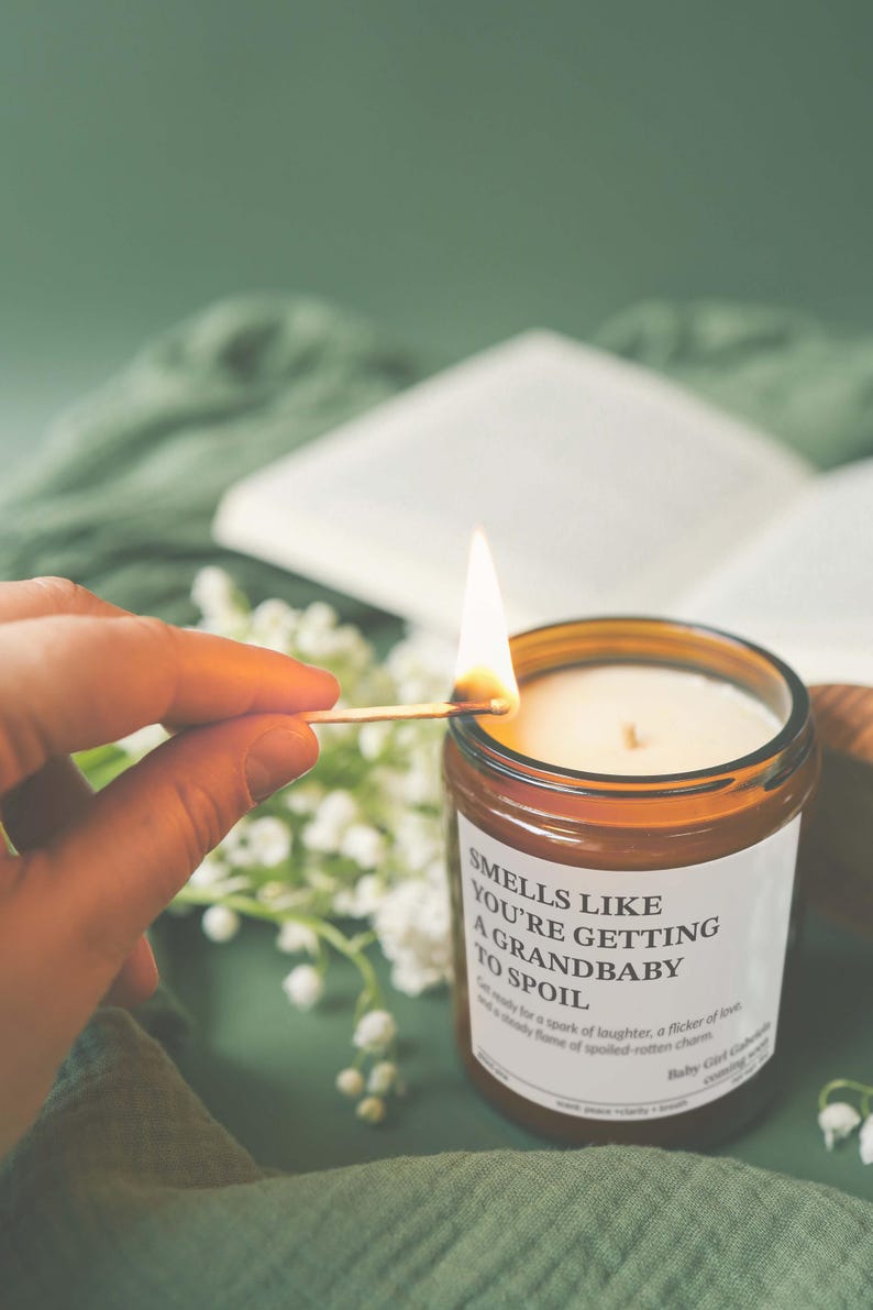 A hand is lighting a candle with a match, and the candle is labeled with the text "Smells like you're getting to spoil a grandbaby".
