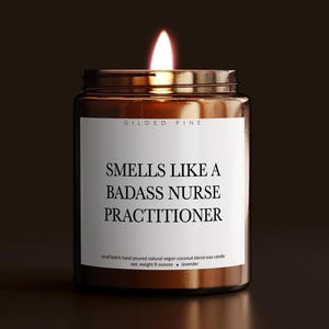 May include: A lit, brown glass jar candle with a white label. The label reads "SMELLS LIKE A BADASS NURSE PRACTITIONER." The candle is a small batch, hand-poured, natural, vegan coconut blend wax candle, net weight 8 ounces. The scent is lavender.