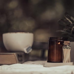 a table with various objects, including a brown candle, a white bowl, a brown jar, a brown bottle, a brown vase, and a brown book. The objects are arranged in a casual and relaxed setting, with the focus on the table and its contents.