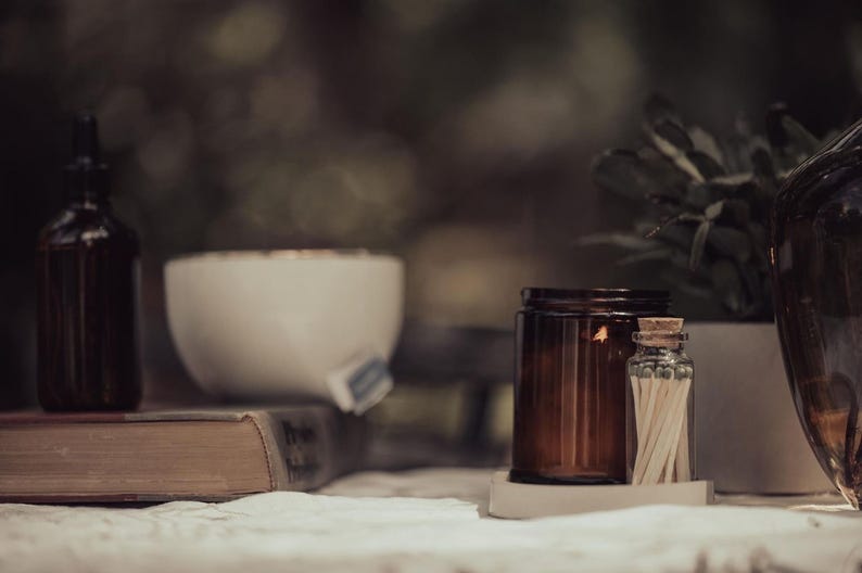 a table with various objects, including a brown candle, a white bowl, a brown jar, a brown bottle, a brown vase, and a brown book. The objects are arranged in a casual and relaxed setting, with the focus on the table and its contents.