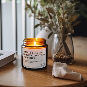 a candle labeled "Smells Like We Survived 16 Years Together" on a wooden table, with a vase of greenery and a book in the background.