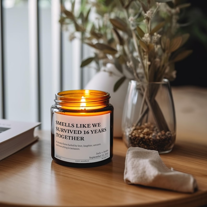 a candle labeled "Smells Like We Survived 16 Years Together" on a wooden table, with a vase of greenery and a book in the background.