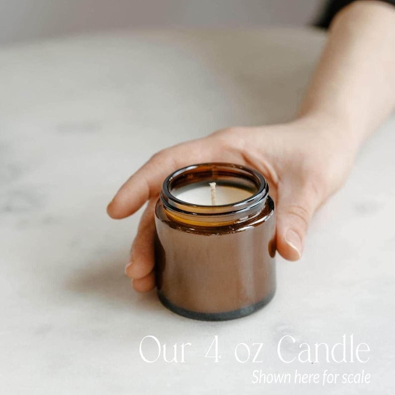 A hand holding a brown glass jar with a white candle inside, placed on a white surface.