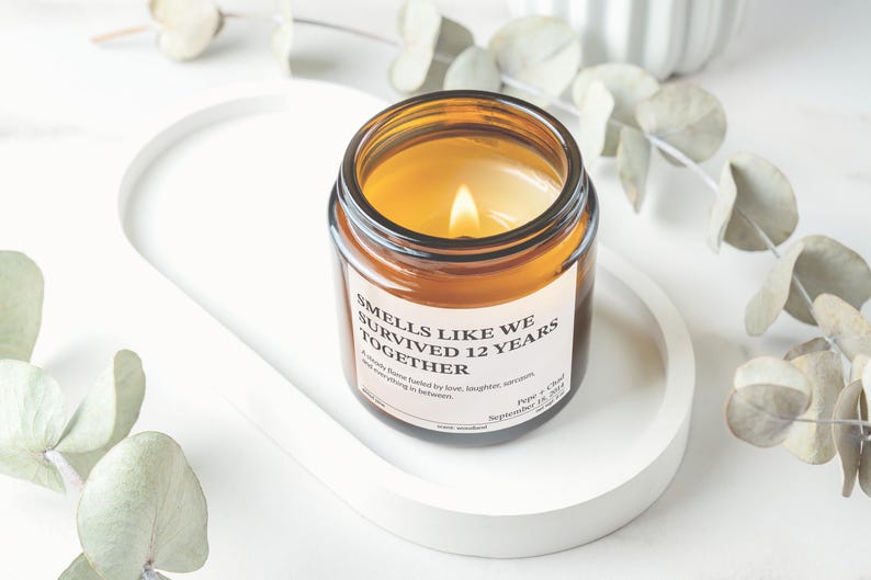A brown glass jar with a white label sits on a white tray, surrounded by eucalyptus leaves and other greenery.