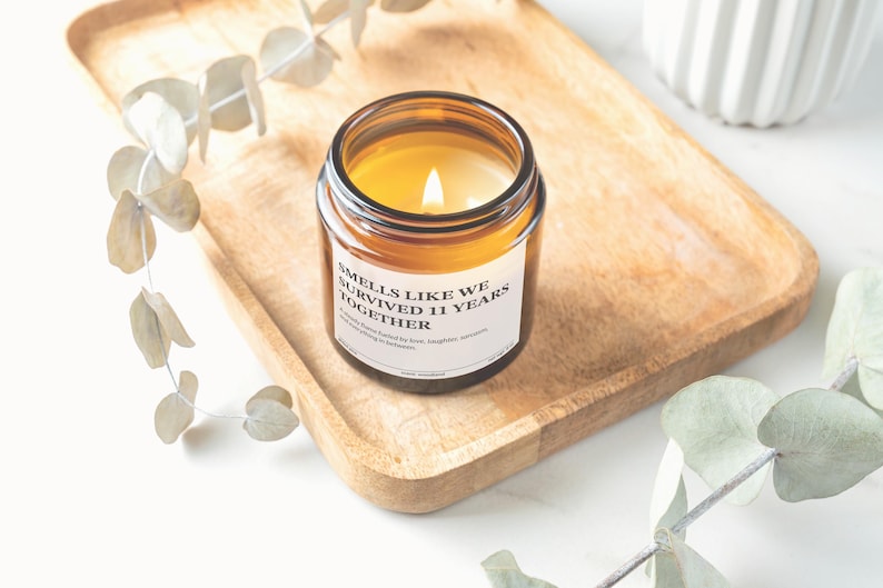 A brown glass candle with a white label sits on a wooden tray, surrounded by greenery and a white vase.