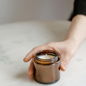 A hand holding a brown glass jar with a white candle inside, placed on a white surface.